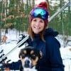 A woman wearing a hat and goggles is holding a dog in the snow.