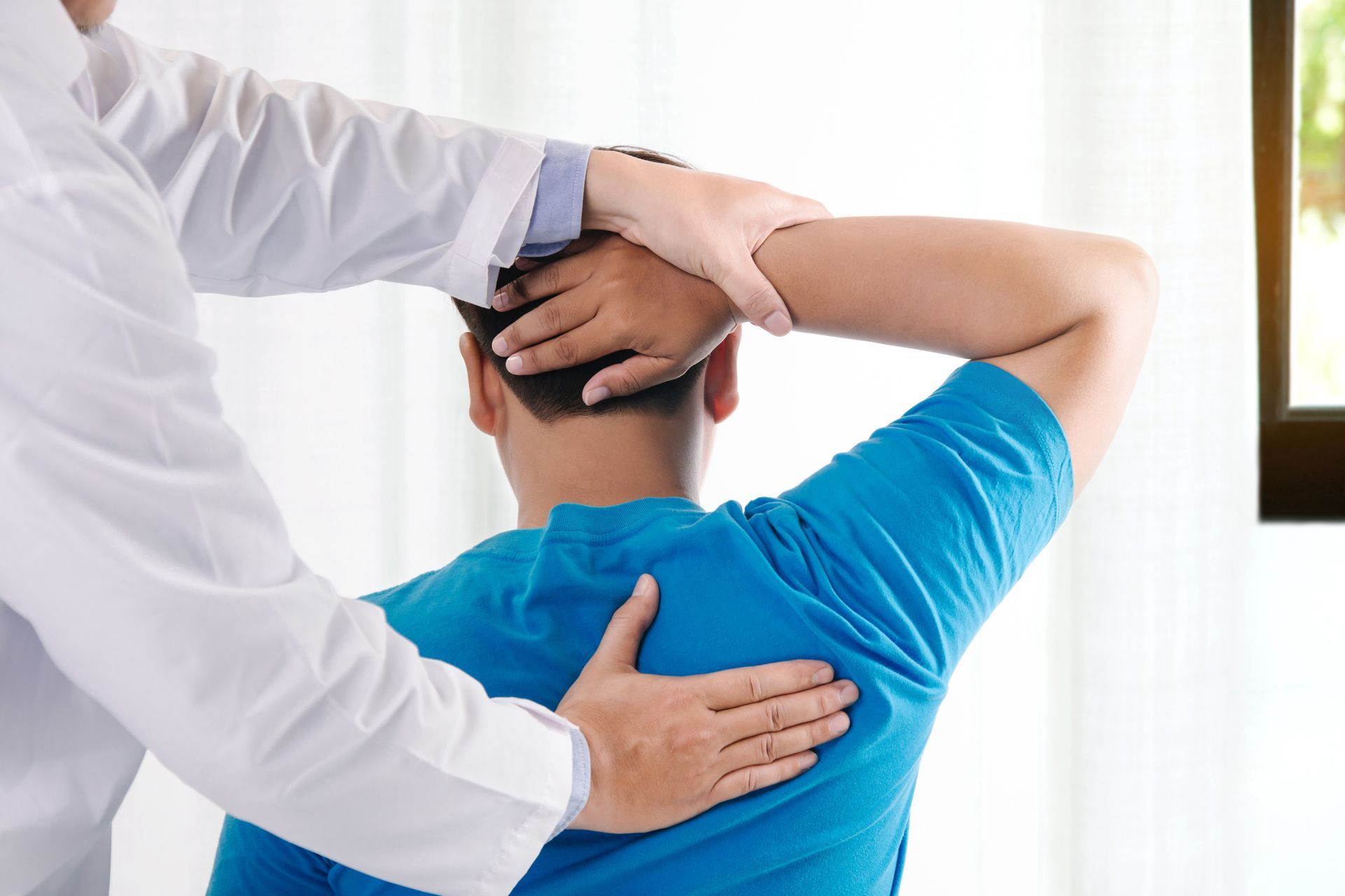 A healthcare professional in a white coat performs a physical examination on a patient's back and neck in a bright room.