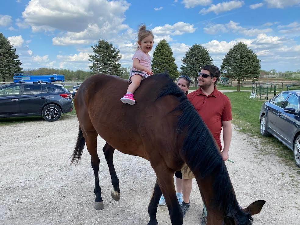 Child riding a brown horse while an adult stands beside it in a grassy parking area under a cloudy sky