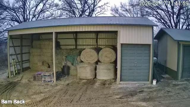 Open-front barn filled with hay bales and farm equipment, with a metal shed door on the right.