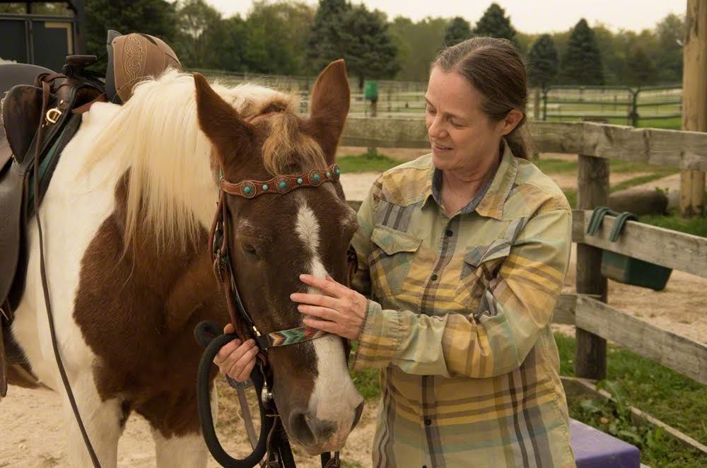 Woman petting a brown-and-white horse in a fenced pasture outdoors