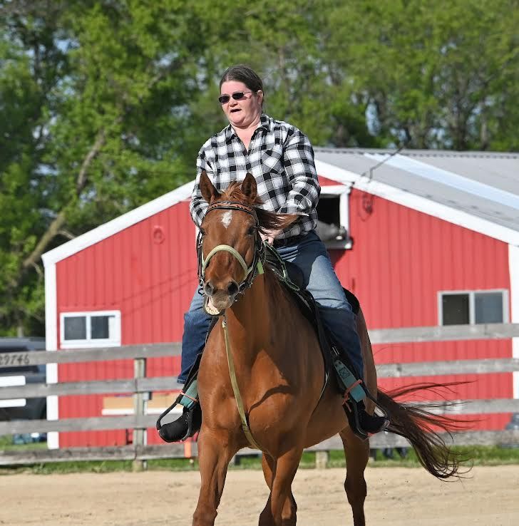 Rider on a brown horse in front of a red barn, wearing a plaid shirt and sunglasses.
