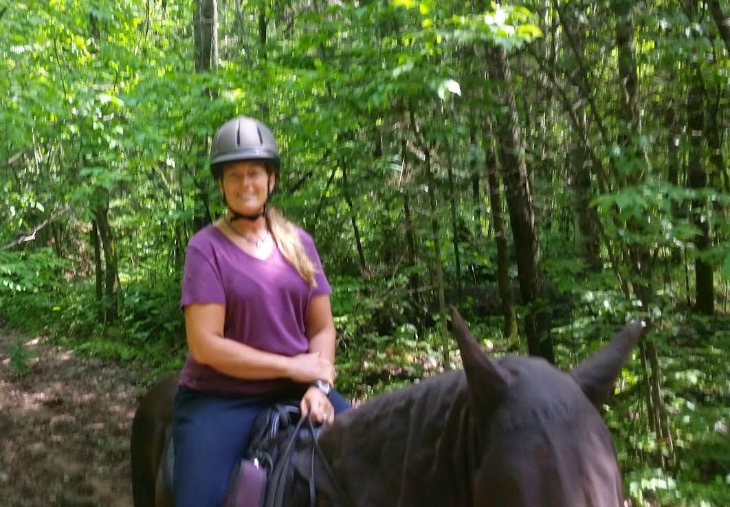 Person riding a horse on a forest trail, wearing a helmet and purple shirt.