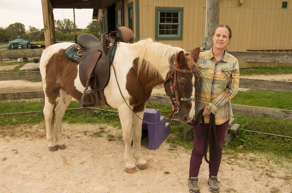 Brown-and-white horse saddled beside a person standing outside a barn