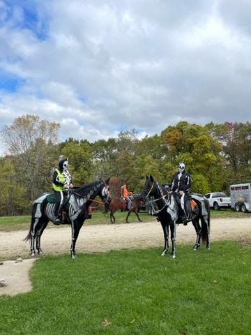 Mounted riders on black horses in a grassy park, with a gravel path, trees, and cloudy sky.