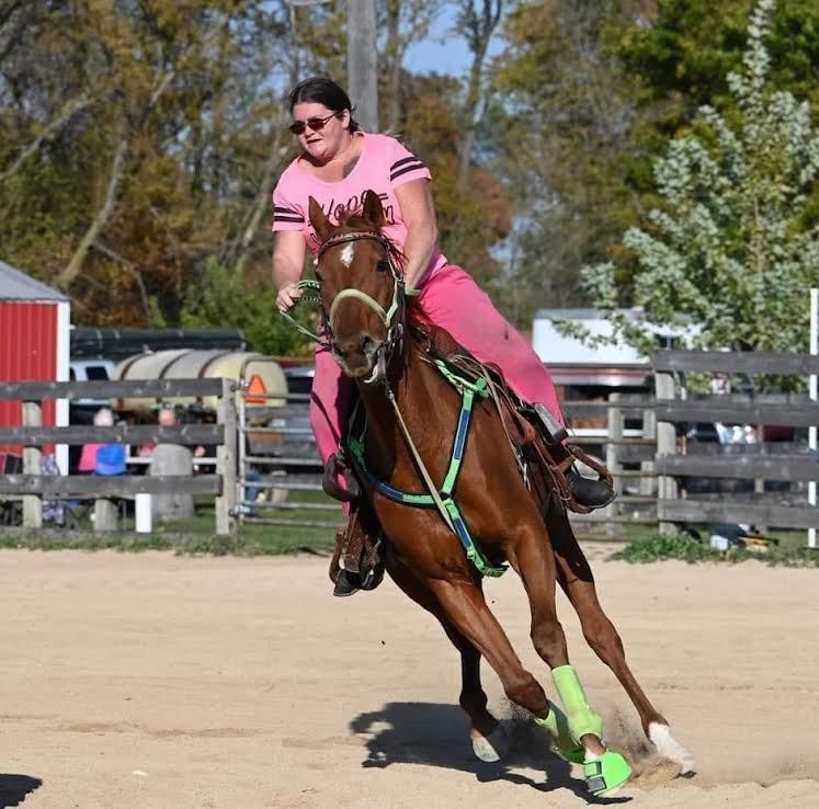 Rider in pink shirt and pants gallops a chestnut horse on a dirt arena, leaning into a turn.