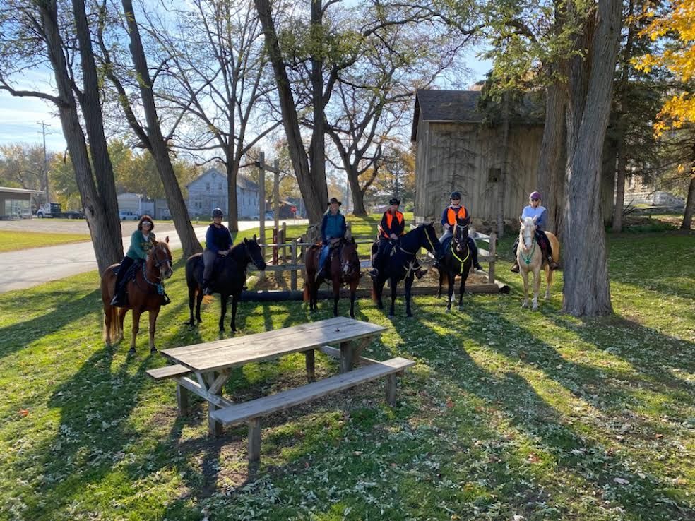 People on horseback lined up in a sunny park beside a picnic table and trees