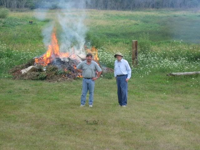 Two people stand in a grassy field beside a smoking bonfire.