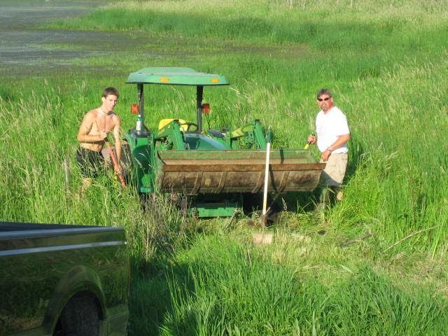 Two people with a green tractor in a grassy field, one standing beside the front loader.