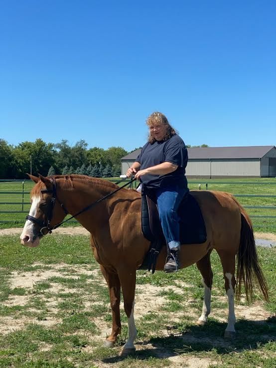 Person riding a brown horse in a sunny field with a barn in the background