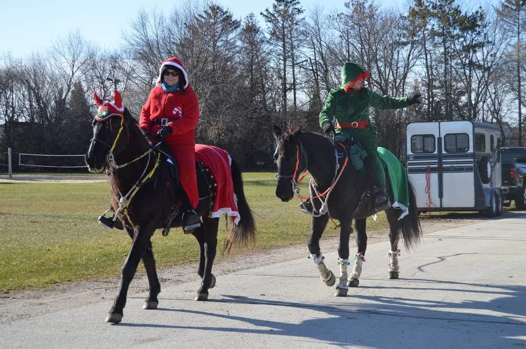 Two people in red and green outfits ride horses on a sunny path near a barn.