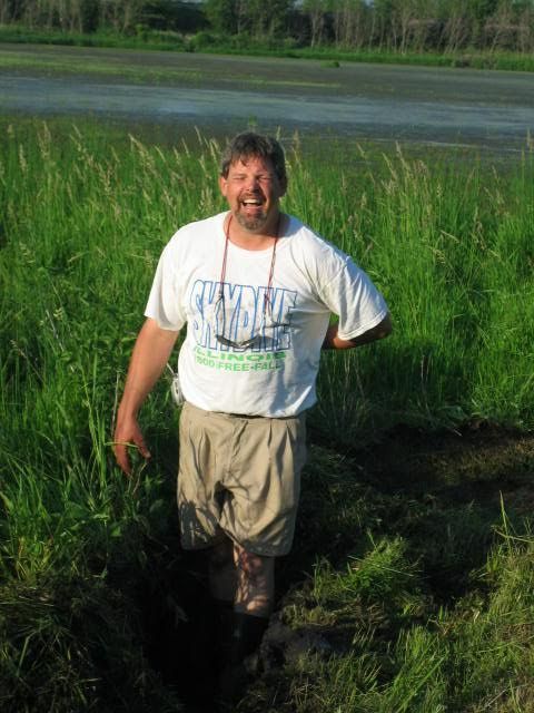 Man standing in tall grass beside a wetland, wearing a white T-shirt and khaki shorts, smiling at the camera