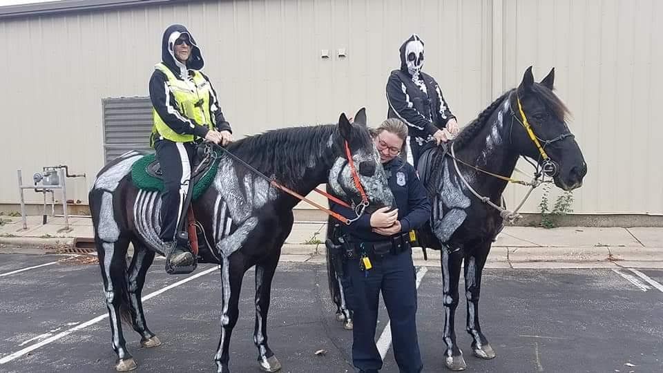 Two saddled horses with riders and a handler standing in front of a building.