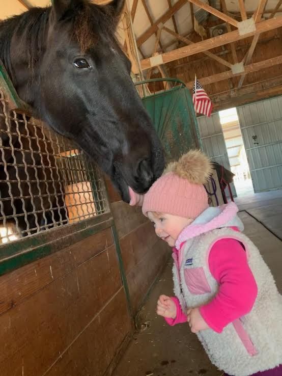 Horse nuzzling a child in a stable, with the child wearing a pink hat and vest