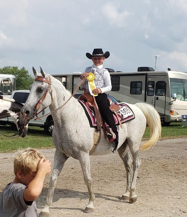 Woman in cowboy hat on white horse at an outdoor rodeo, holding a yellow ribbon.
