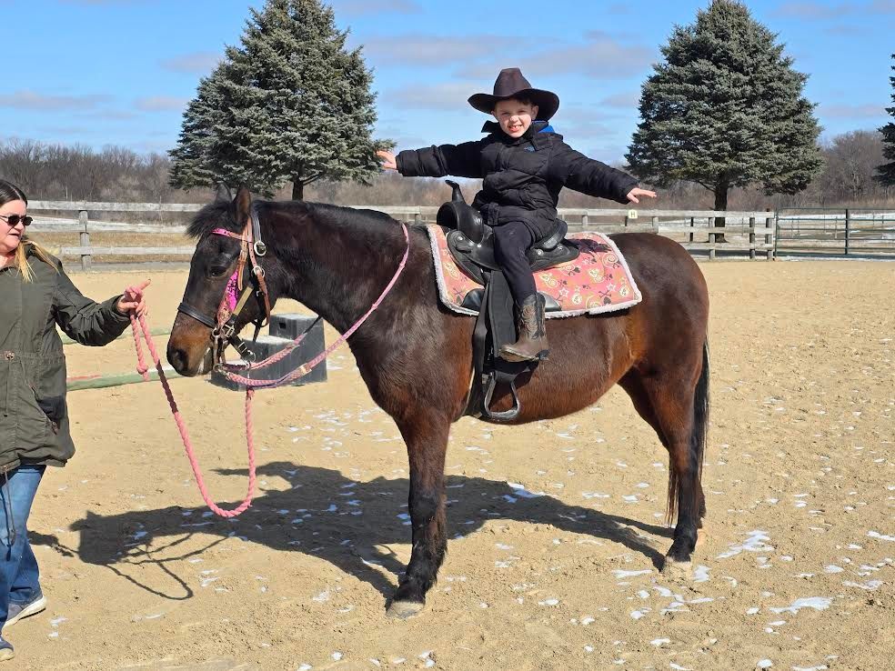 Person in a cowboy hat riding a dark horse while another person holds the reins in a paddock.