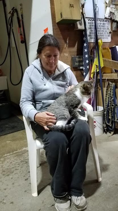 Woman sitting on a white chair with a gray-and-white cat on her lap in a cluttered indoor space