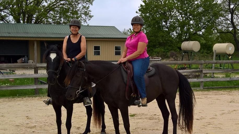 Two riders on black horses in an outdoor riding arena, one in a pink shirt and one in black.