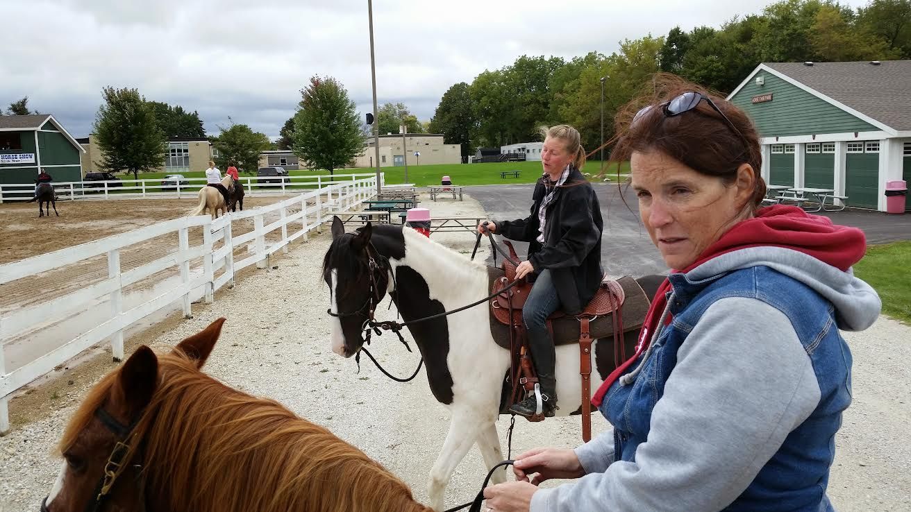 People riding horses in an outdoor arena, with a woman in a blue hoodie in the foreground.