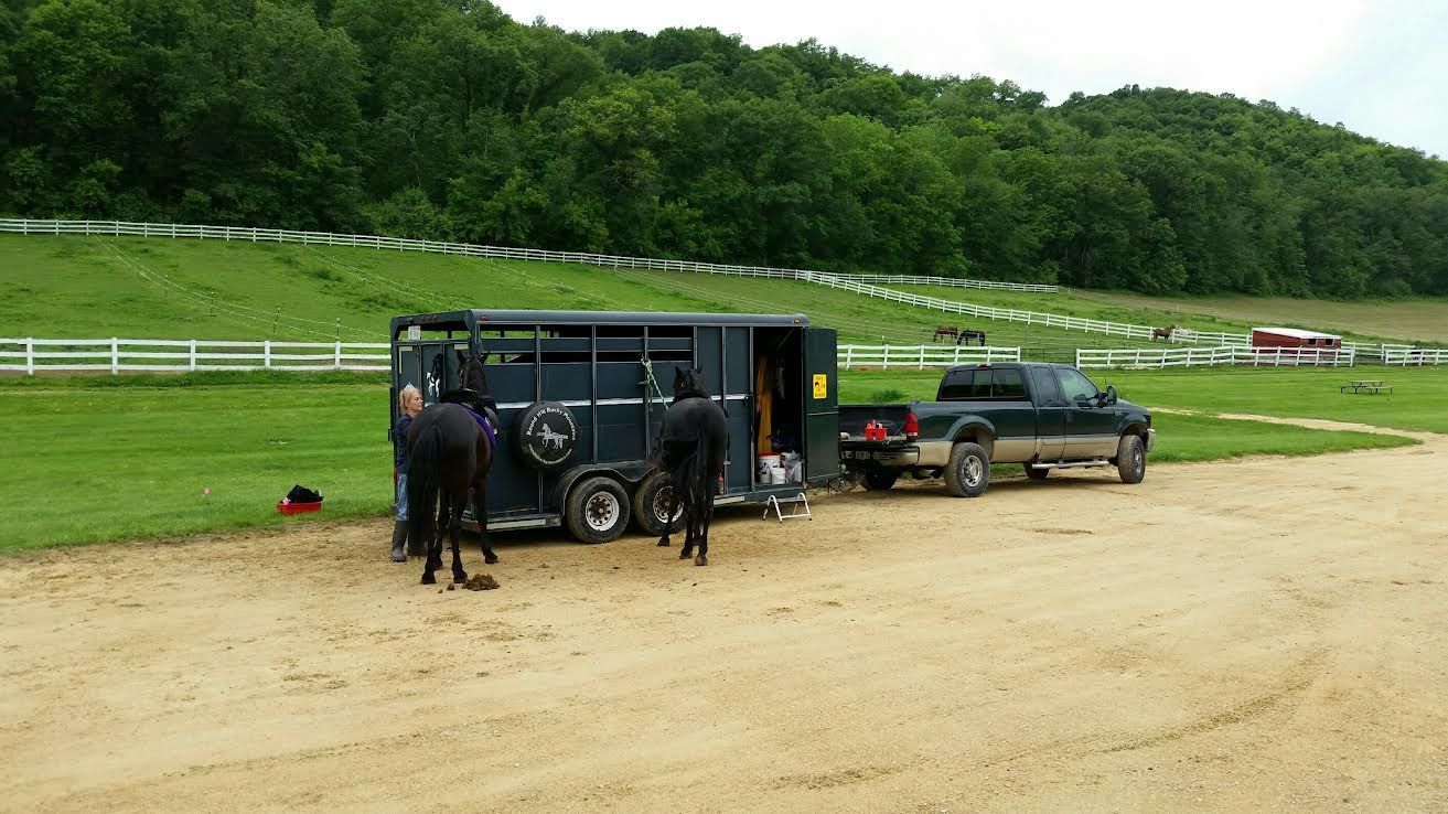 People unloading gear from a black trailer beside a parked car on a dirt road near grassy hills