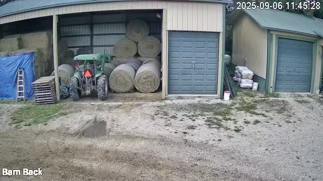Barn with stacked hay bales, a tractor, and closed blue doors on a dirt lot.