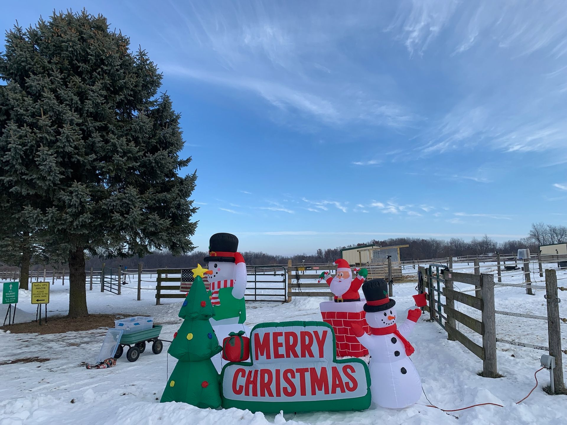 Inflatable Christmas decorations in snowy yard with Merry Christmas sign and decorated fence.