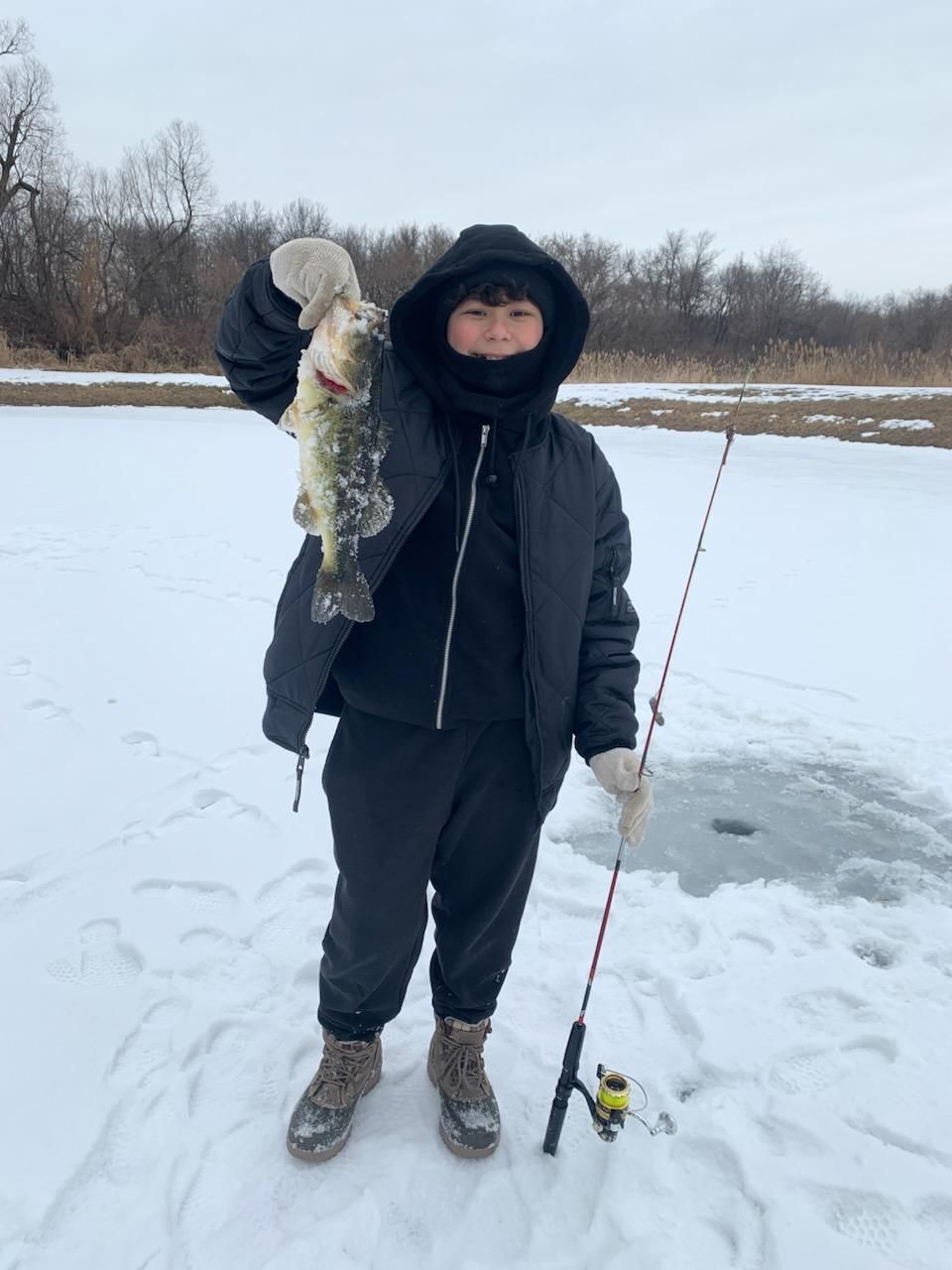 Person in winter clothing holding a fish and a fishing rod on a snowy frozen lake.