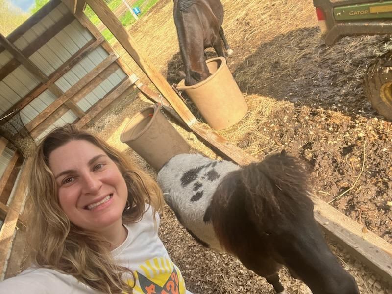 Smiling person taking a selfie with two horses in a stable area outdoors