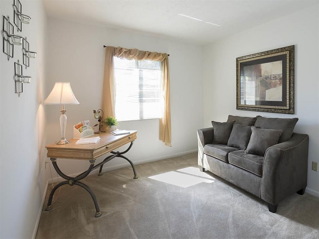 Sunlit living area with a gray sofa, wooden desk, wall art, and a window with beige curtains at Shadow Hill, offering apartments in Sharonville, OH.