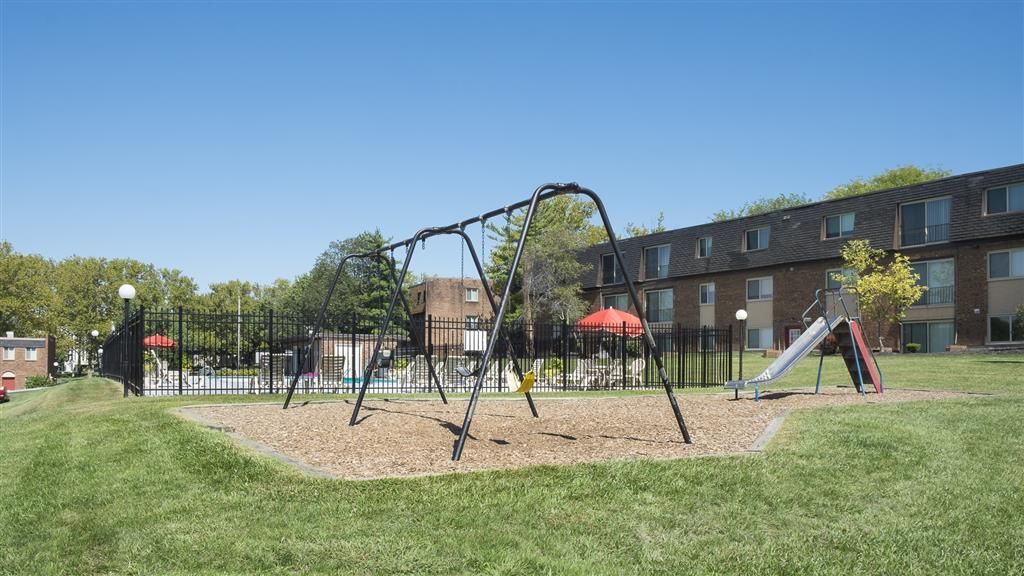 Playground with swings and a slide at an apartment community, with a fenced pool area visible in the background at Shadow Hill, offering apartments near Cincinnati,OH.