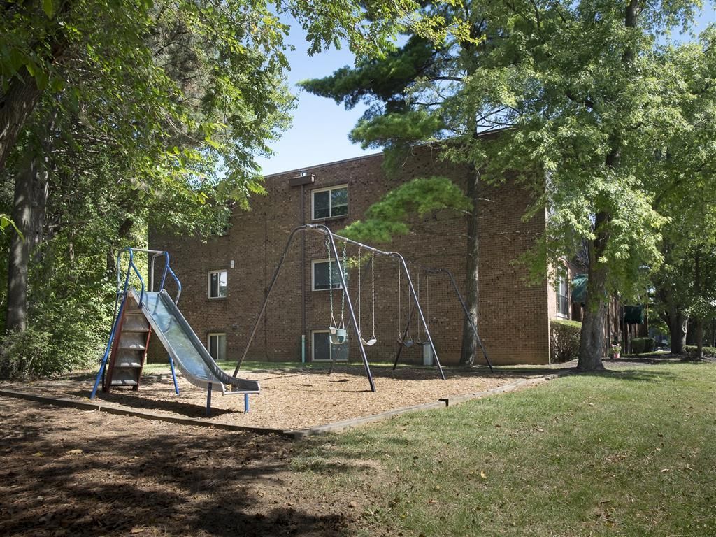 Playground with slide and swings beside a brick apartment building under trees at Shadow Hill, offering pet-friendly apartments near Cincinnati,OH.