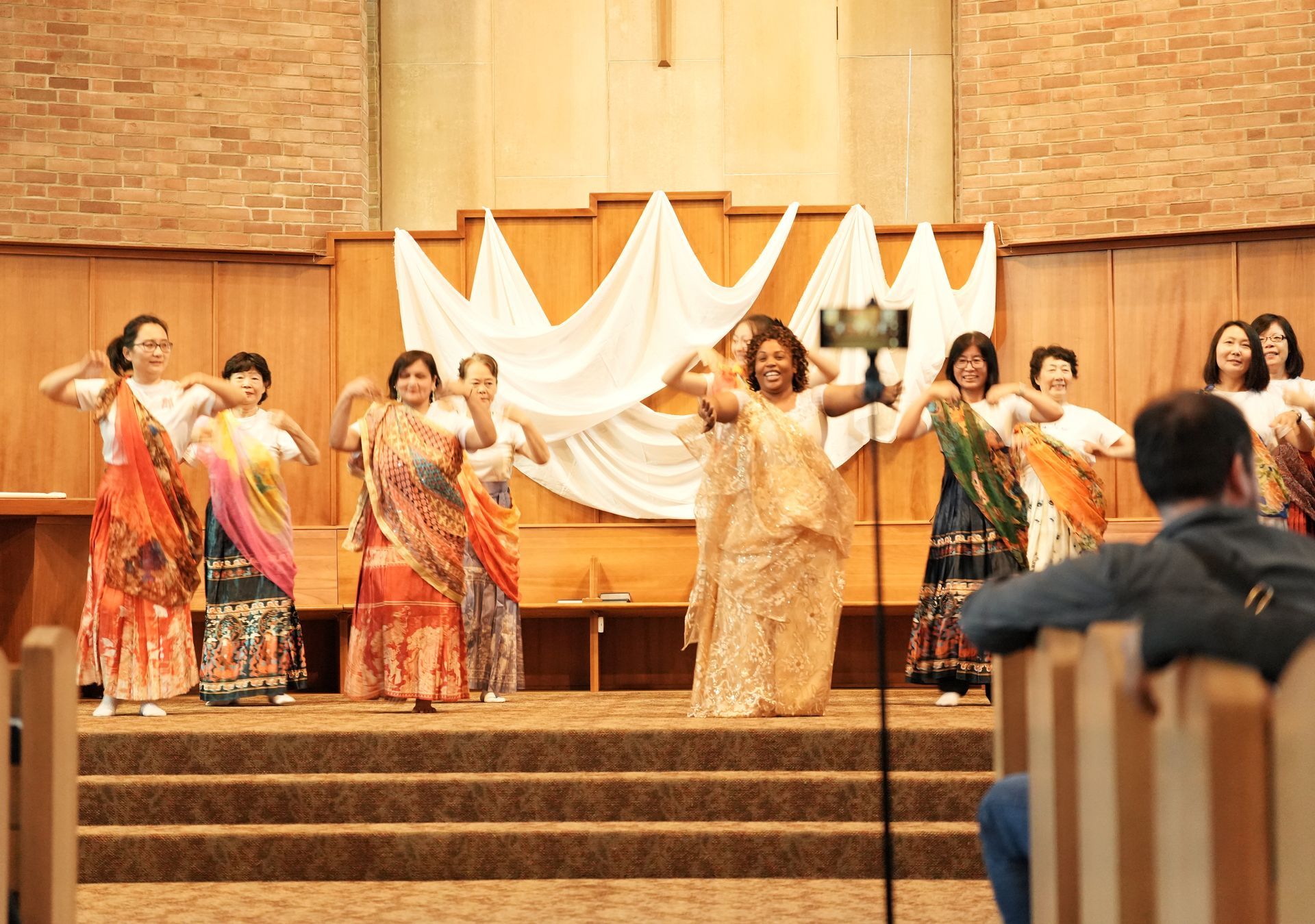 Group in colorful traditional outfits posing indoors at a celebration, with raised hands and decorated backdrop.