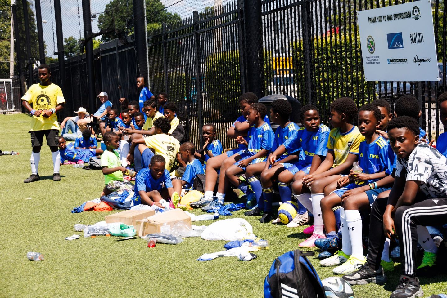 Children in blue shirts sitting on grass at an outdoor sports event