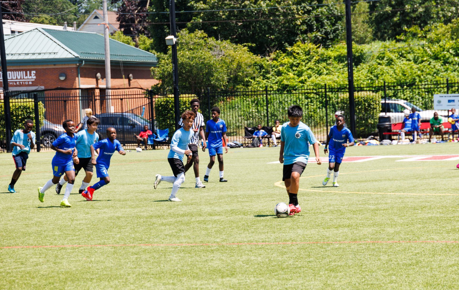 Youth soccer players in blue compete on a grassy field near a building and trees.