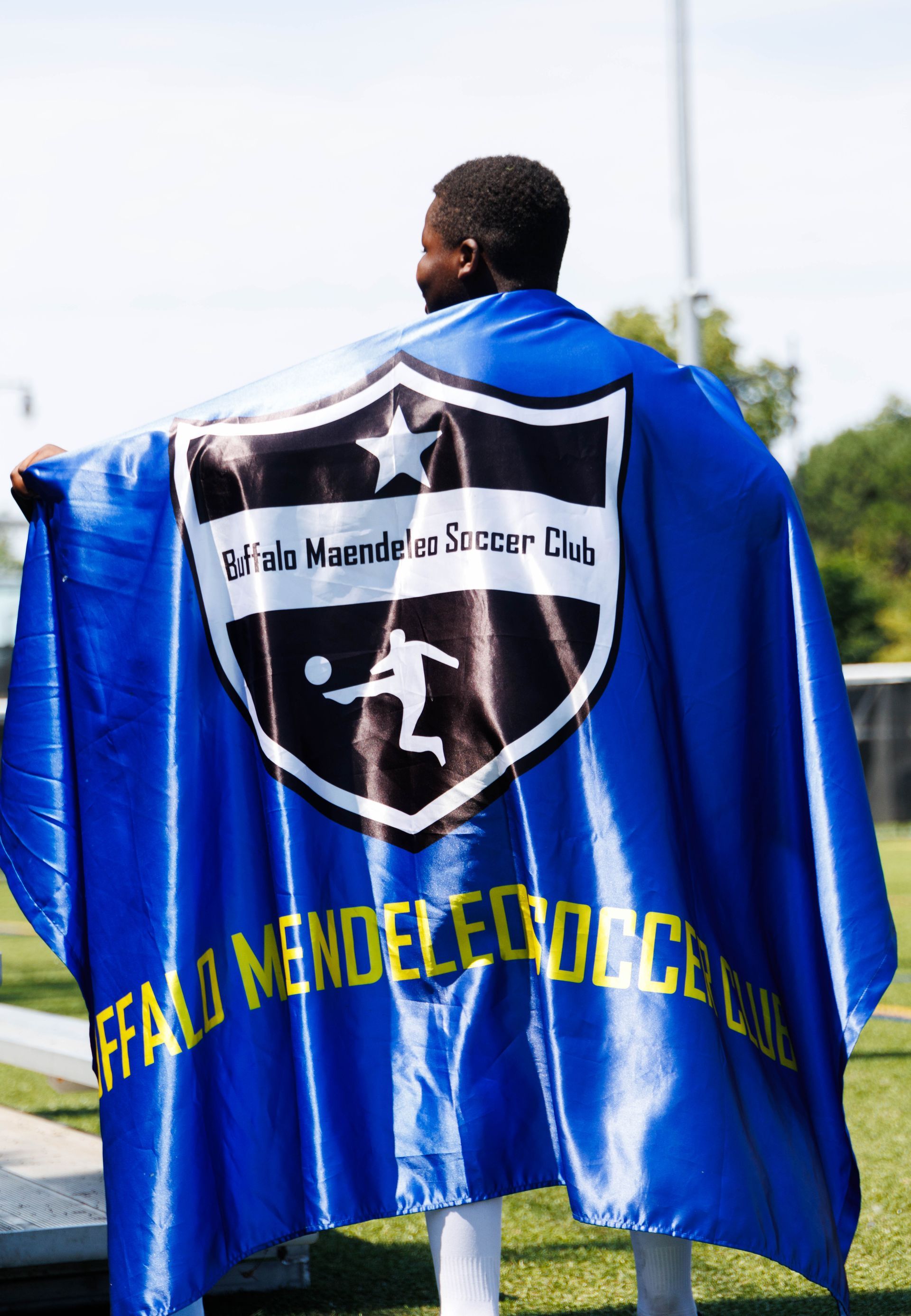 Person draped in blue cape with soccer club crest and yellow lettering outdoors