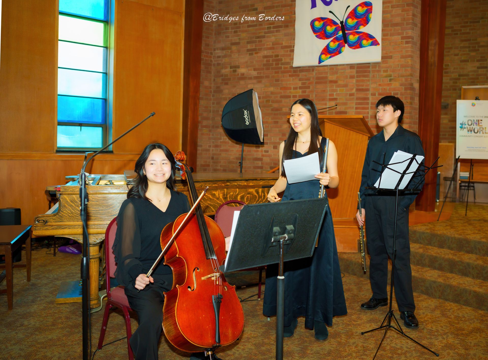 Three musicians rehearsing in a brick hall, with a cello, microphone, and music stands.