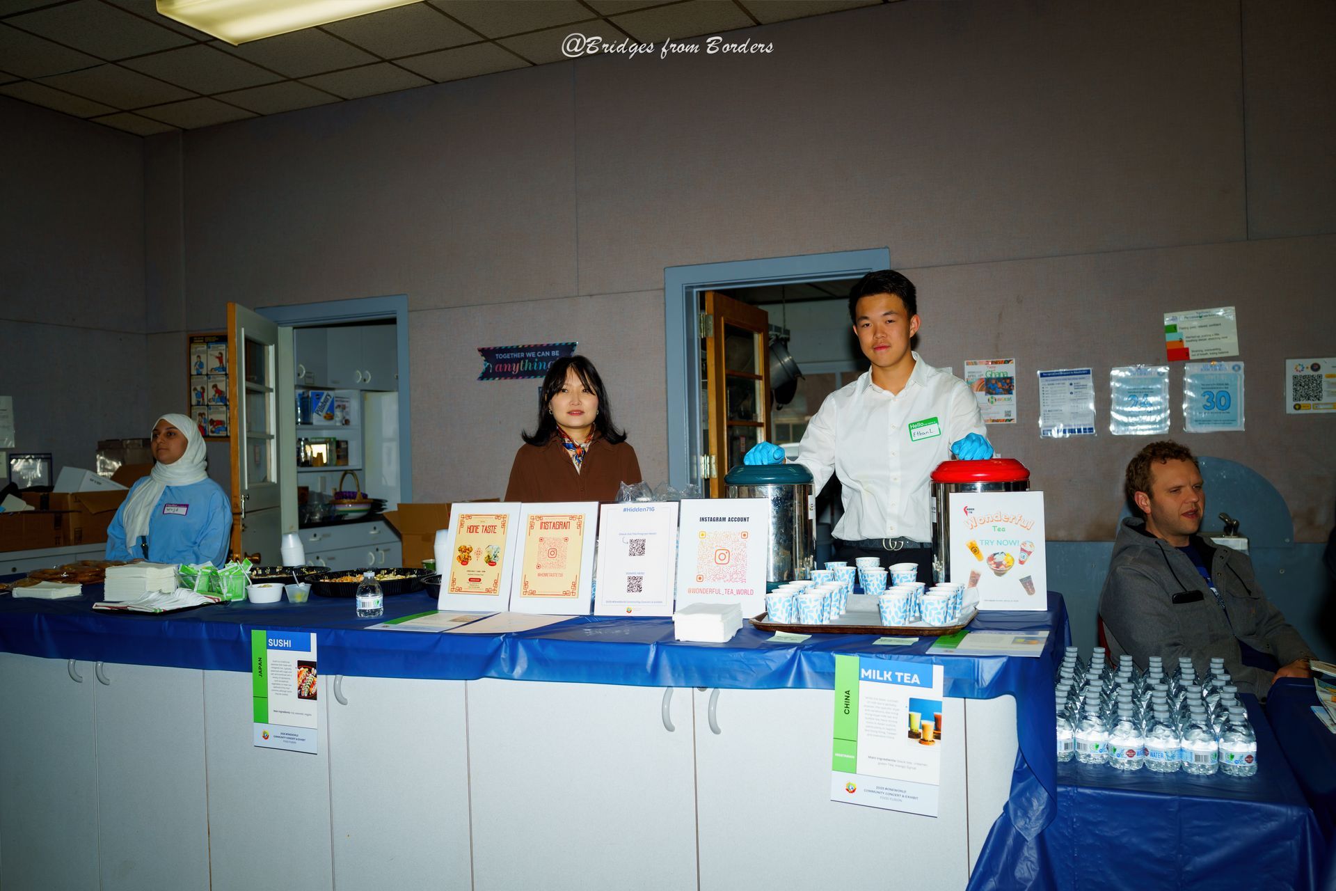 People at a blue-clothed beverage table with bottled drinks and display signs in an indoor hall