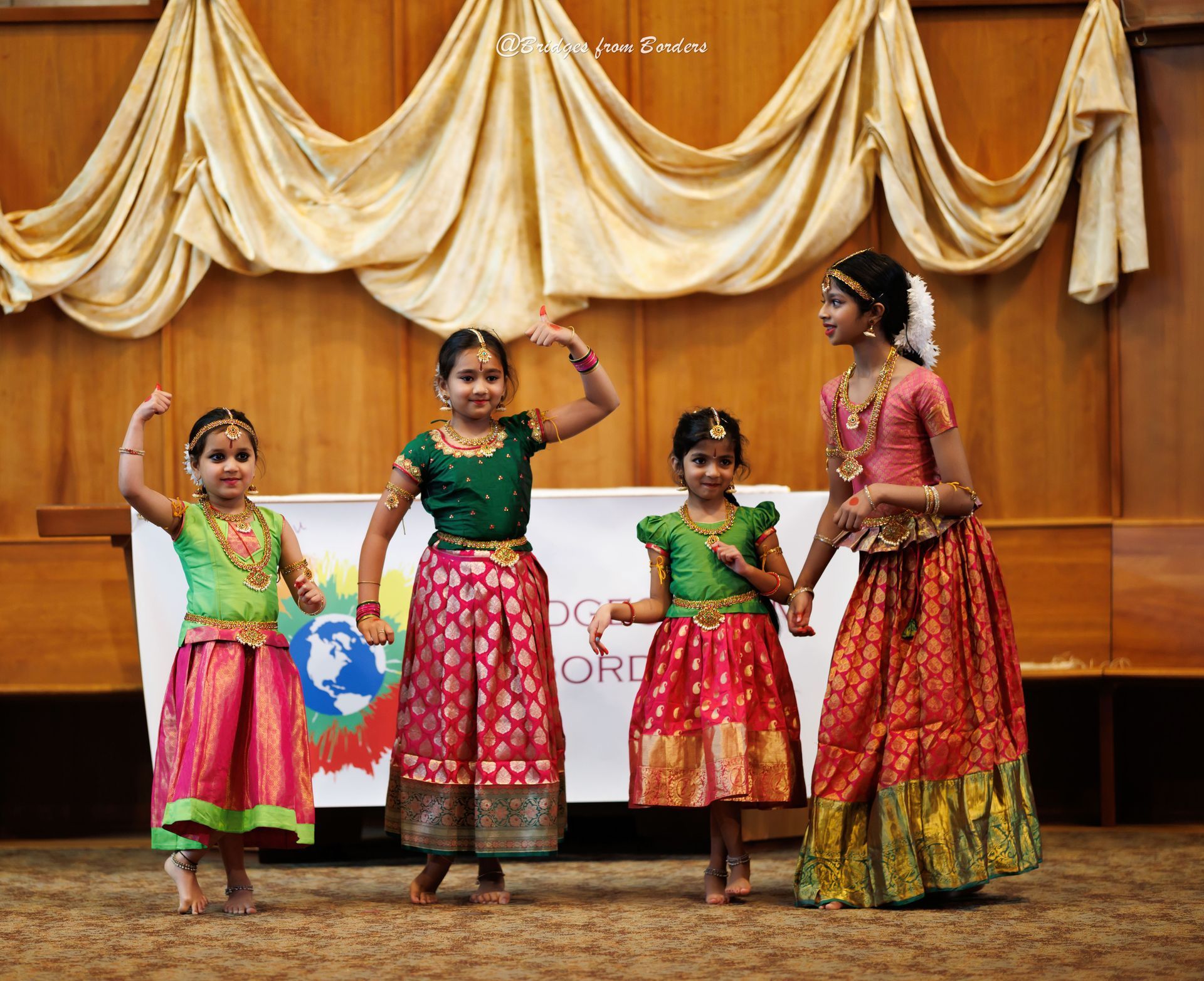Four children in colorful traditional dance costumes posing on a stage with draped curtains.