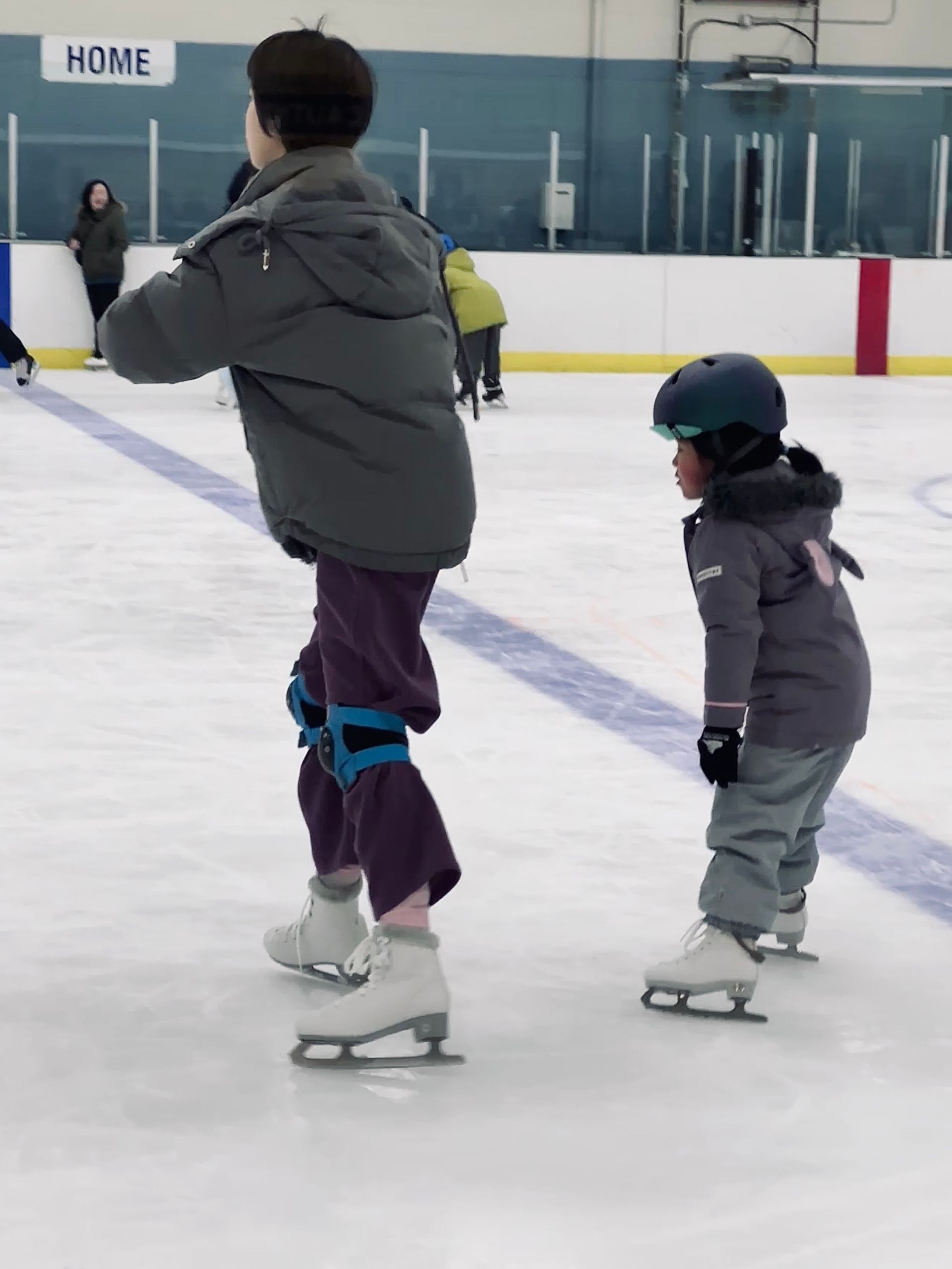 Two people ice skating indoors, one helping the other on a rink.