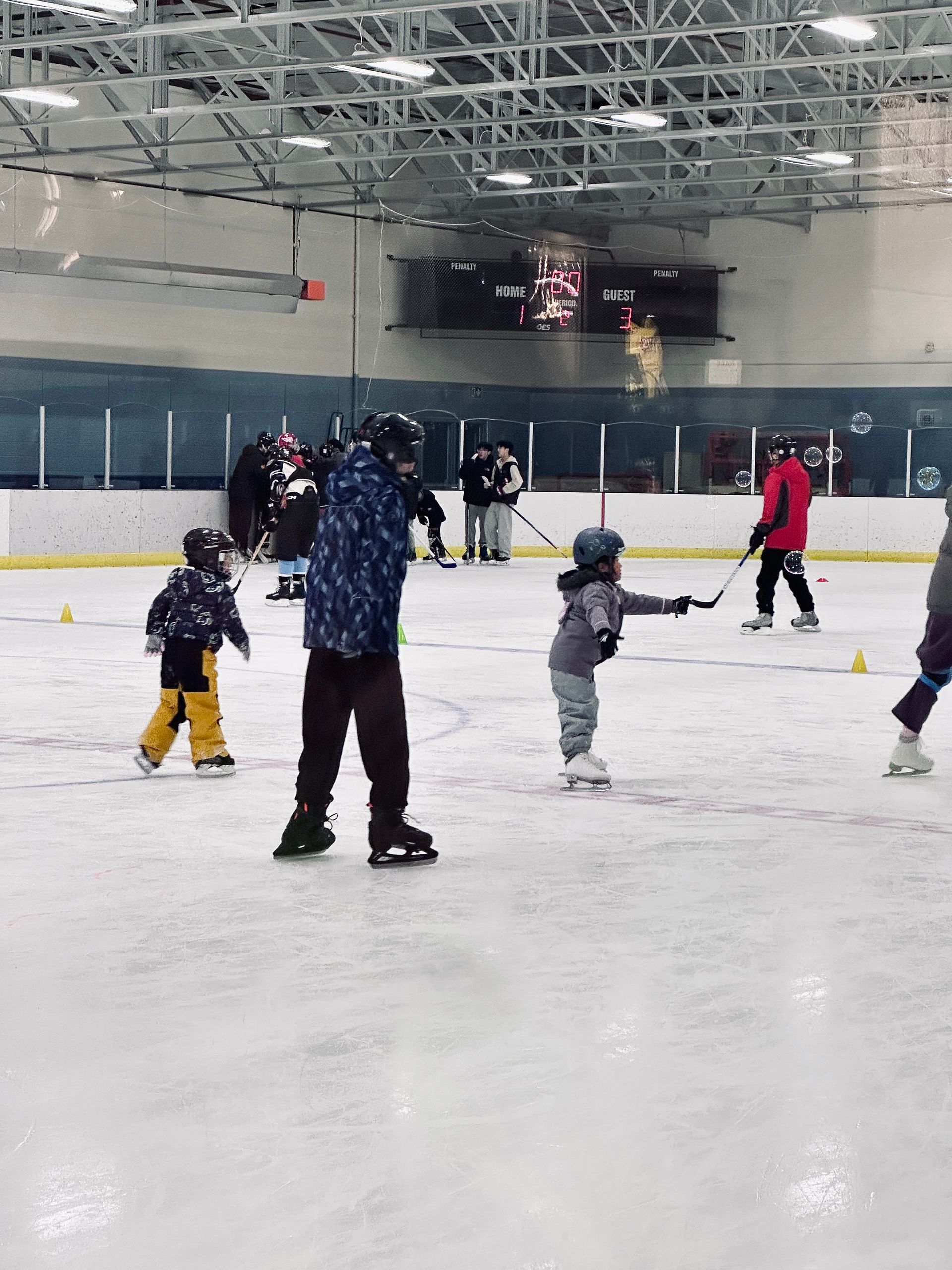 Indoor ice rink with children and adults skating, viewed from behind.