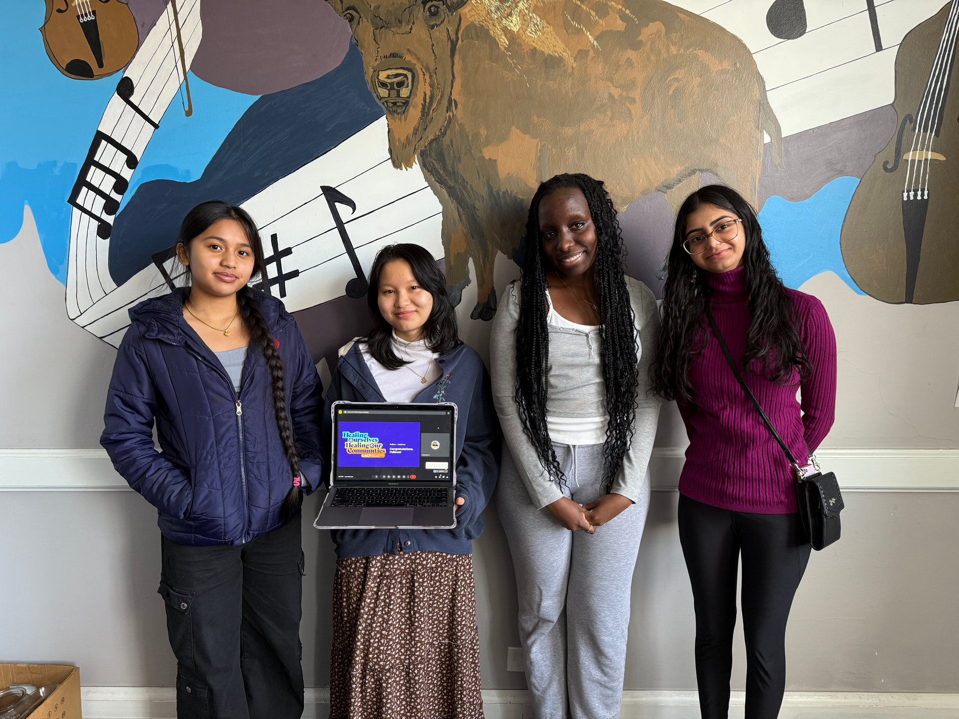 Four people stand by a laptop display in front of a colorful mural, smiling at the camera.