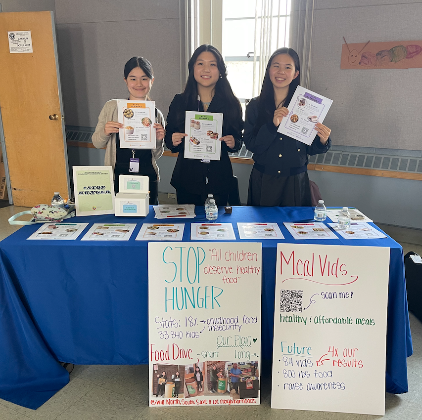 Three people behind a blue table display posters about food insecurity at an indoor event.