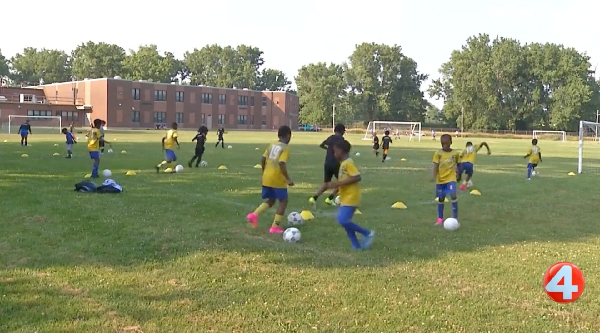 Children practicing soccer on a grassy field with cones and a brick building in the background