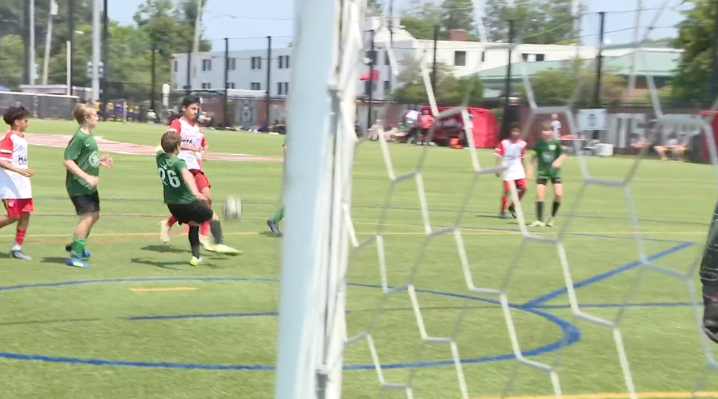 Soccer players near the goal on a grassy field, with a goalkeeper and net in the foreground.