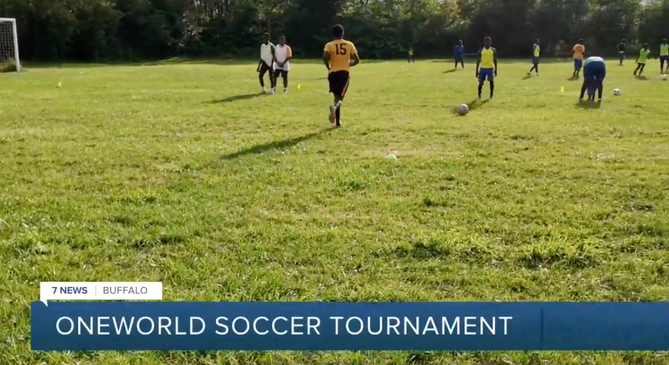 Soccer players on a grassy field during the OneWorld Soccer Tournament, with a scoreboard banner at the bottom.