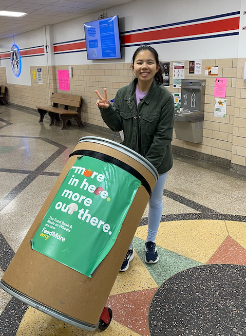 Smiling person posing beside a large green recycling bin in a school hallway