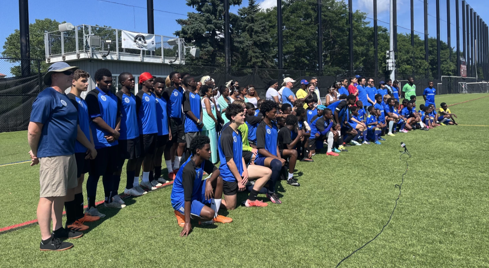 Large group in blue sports uniforms posing on a grassy field beside a netted court