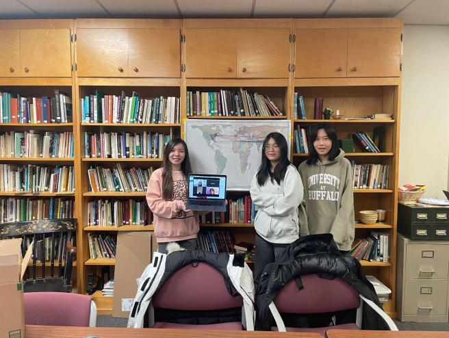 Three people posing in a library with a laptop on a chair in front of bookshelves.