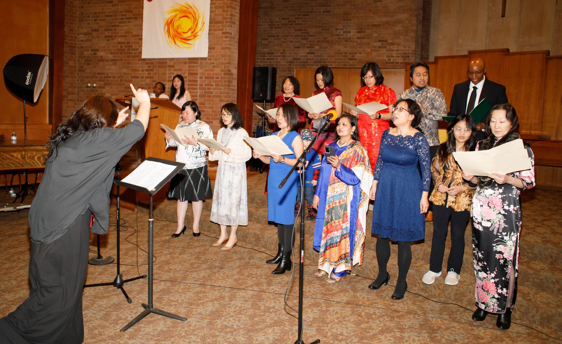 Choir singing in a brick-walled hall, with a conductor leading and microphones in front