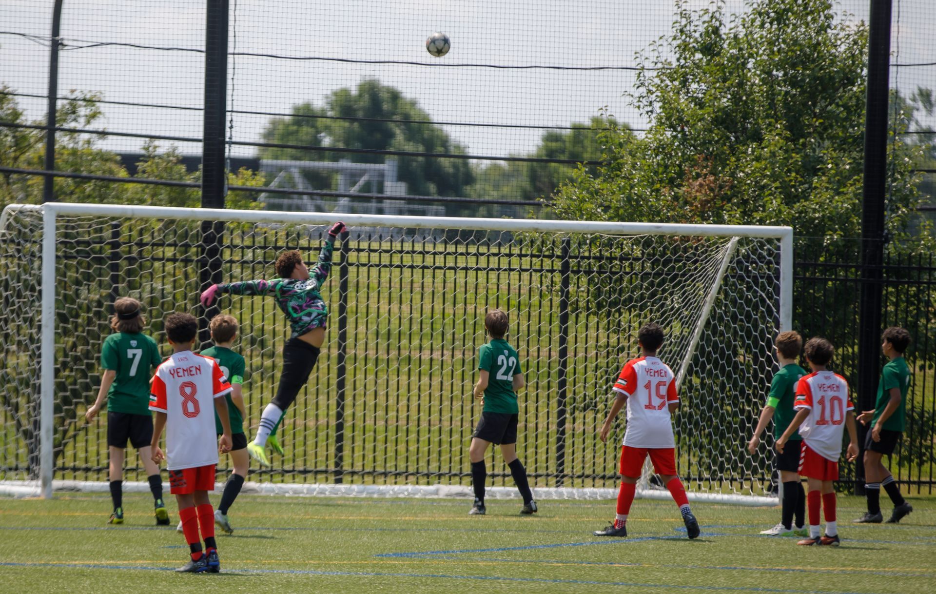 Youth soccer players near the goal as a goalkeeper jumps to catch a high ball on a sunny field